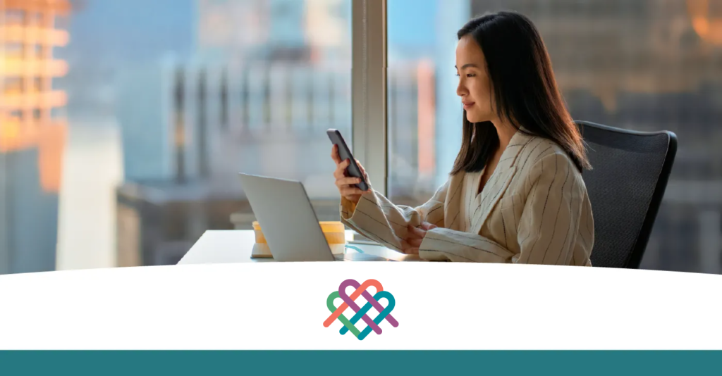 Photo of a woman in an office with a laptop on the desk. She is looking at the phone in her hand.